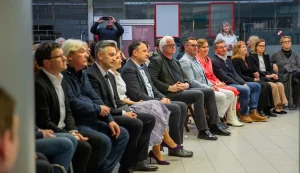 Un gruppo di persone è seduto in fila e ascolta una presentazione durante un evento che si tiene in un locale al chiuso.