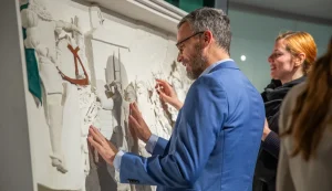 Un uomo in una galleria osserva con le mani un'opera d'arte murale a rilievo con figure in rilievo.