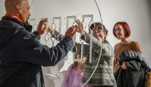 Due visitatori in una galleria esplorano con le mani un'installazione artistica tattile composta da elementi sospesi e testurizzati, pensata per essere percepita attraverso il tatto.