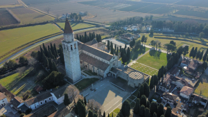 Veduta aerea della Basilica di Aquileia con il caratteristico campanile, circondata da filari di alberi e dal paesaggio pianeggiante circostante.