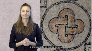 Un’interprete sta in piedi davanti alla telecamera e presenta il contenuto; accanto a lei è visibile un motivo a mosaico con un ornamento intrecciato.
