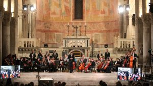 Orchestra con direttore si esibisce sul palco all’interno di una chiesa con alte colonne e affreschi; i musicisti sono seduti in semicerchio con strumenti ad arco e altri strumenti, sullo sfondo si trova l’altare.
