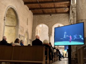 Visitatori seduti su panche di legno all’interno di una chiesa antica con arcate guardano un grande schermo su cui è mostrata una ballerina all’aperto.