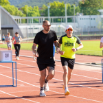 Diversi corridori corrono su una pista di atletica; lungo il percorso sono presenti scatole con codici QR per le donazioni.