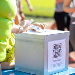 Una persona inserisce un biglietto in una scatola per donazioni; sulla scatola è presente un codice QR per donazioni via SMS.
