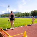 Un adulto e un bambino bendati camminano su una pista di atletica con bastone bianco, superando ostacoli segnalati da coni.