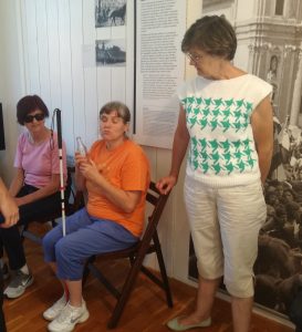 Tre donne in un museo; quella al centro tiene davanti a sé una bottiglia di Coca-Cola e ha un bastone bianco.