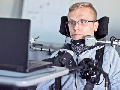 Una persona utilizza una carrozzina, interruttori e altri dispositivi assistivi per usare il computer.