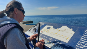 Una persona con bastone bianco utilizza una mappa tattile con indicazioni in rilievo sul lungomare per orientarsi nello spazio.
