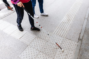 Una persona con bastone bianco rileva una superficie tattile di attenzione e un percorso tattile.