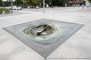 Fontana in bronzo a livello del suolo al centro di una piazza in pietra, dalla quale sgorga l’acqua.