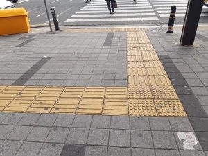 Segnaletica tattile a pavimento con percorsi guida e superfici di attenzione che conducono verso l’attraversamento pedonale.